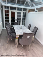 A gray marble dining table with six gray velvet chairs with a lion knocker back design, placed in a room with a window and a decorative object in the background.