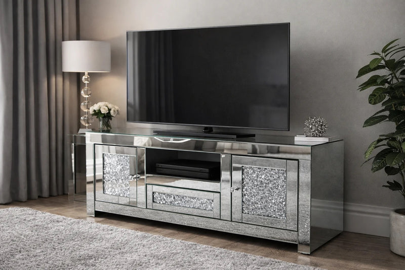 Silver mirrored TV stand with a television mounted on a gray wall. The Roma TV Unit is designed to complement contemporary, glam, and luxury interiors. Its mirrored surfaces and crystal-style accents add brightness and depth to your room, making it an eye-catching centrepiece whether styled alone or paired with matching Roma furniture.
Sizes Available in two sizes to suit your space and TV setup of your 

120 cm (W) x 45 cm (D) x 55 cm (H)
150 cm (W) x 45 cm (D) x 55 cm (H)

