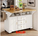 A white kitchen island cart with a light wood countertop, shown in a bright kitchen setting. The cart has three drawers with small gold knobs, two cabinet doors with gold handles, open shelving, and black caster wheels.
