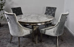 A round dining table with a gray marble-patterned top and a metallic silver base, surrounded by four matching gray velvet armchairs with silver nailhead trim and ring pulls, set in a room with gray carpet and light gray walls.